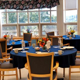 A well-lit dining area with round tables covered in blue tablecloths, set with dishes and floral centerpieces.