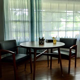 A cozy corner with a round table and two chairs by the window, adorned with sheer curtains and potted plants.