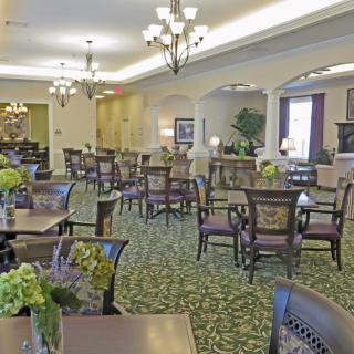 A spacious and elegantly decorated dining room with multiple tables and chairs, chandeliers, and greenery.
