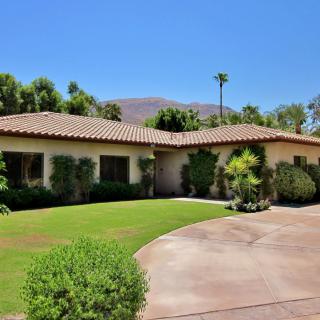 A Spanish-style single-story house with a landscaped garden and palm trees, set against a mountainous backdrop.