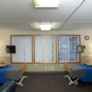 A well-lit hospital room featuring two beds with medical equipment and windows covered by vertical blinds.