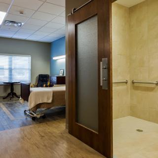 A modern patient room featuring a shower area, a bed, and an office chair by the window.