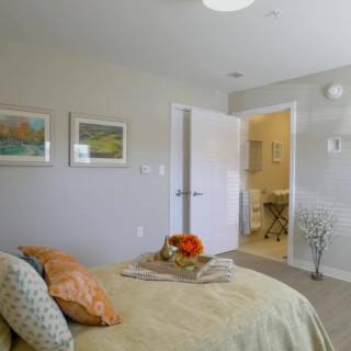 A modern bedroom interior with a neatly made bed, artwork on the walls, and a view into an adjoining bathroom.