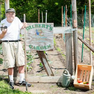 An elderly man stands beside a garden, proudly holding fresh produce with a sign that reads 'Gilbert's Garden.'