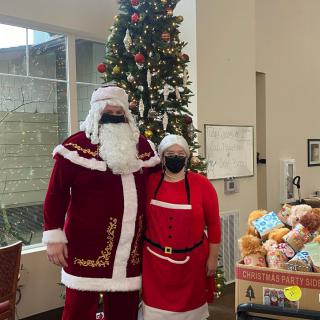 Two people dressed in Santa costumes pose beside a decorated Christmas tree indoors.