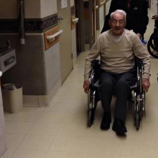 An elderly person in a wheelchair moving through a hospital corridor.