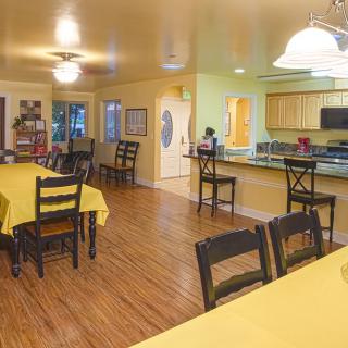 A cozy dining area with wooden floors and multiple tables and chairs, adjacent to a kitchen.