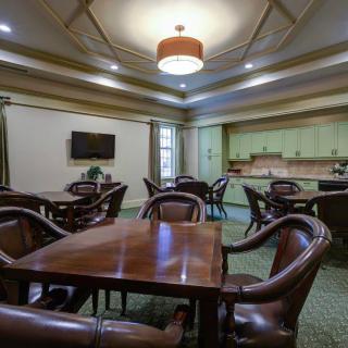 A well-lit dining area featuring several tables and chairs, with a wall-mounted television and a kitchenette in the background.