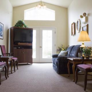 A cozy living room with a sofa, chairs, and a TV stand, accented by decorative artwork and ample natural light from large windows.