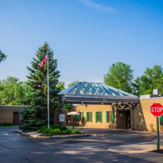 A building entrance with a glass roof, surrounded by trees and featuring a flagpole and a stop sign.