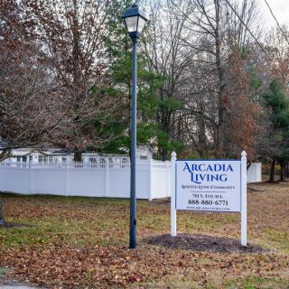 A sign for Arcadia Living stands next to a lamp post, surrounded by a white fence and trees during autumn.