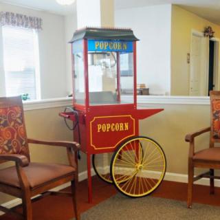 A vintage-style popcorn machine is placed between two chairs in a cozy indoor setting.