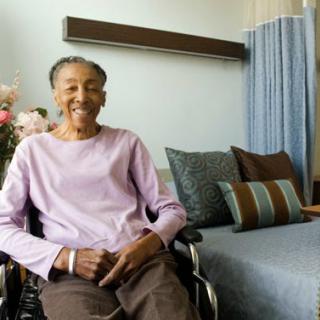 An elderly woman sitting in a wheelchair, smiling warmly in her cozy indoor living space.