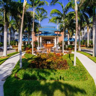 A beautifully landscaped entrance to a residential complex, surrounded by palm trees and vibrant flowers under a clear blue sky.