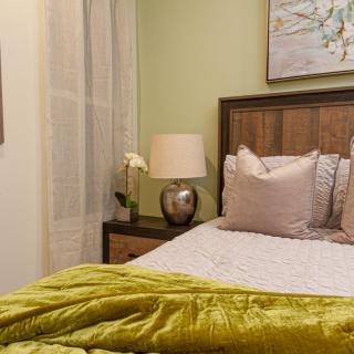 A cozy bedroom with a soft green blanket and decorative pillows, complemented by a stylish lamp and wall art.