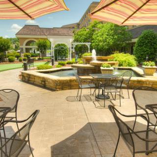 A beautiful outdoor patio area with tables, chairs, and umbrellas surrounding a central fountain, set within a lush garden.
