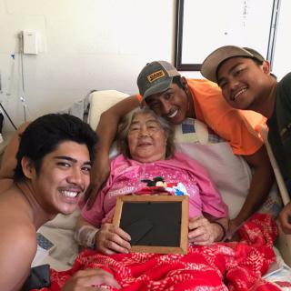 A joyful family gathering around an elderly woman in bed, holding a chalkboard.