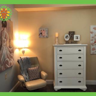 A cozy room with a comfortable chair, a white dresser, and wall art illuminated by a warm lamp.