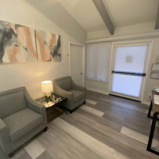 A modern waiting room featuring two gray chairs, a small table with a lamp, and abstract artwork on the wall.