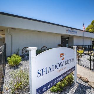 A view of the Shadow Brook building entrance with a sign in front, surrounded by landscaping.