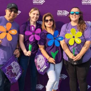 A group of people holding colorful flower props at an outdoor event, all wearing purple shirts and smiling.