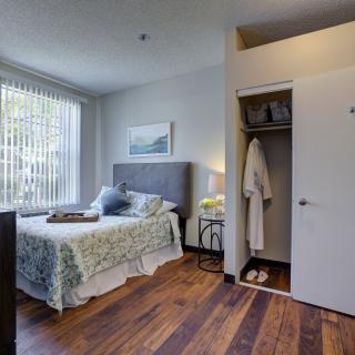 A modern bedroom with hardwood floors, featuring a bed and an open closet displaying a robe and slippers. Natural light filters through the window blinds.