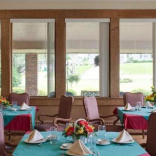 A cozy dining area with neatly set tables, decorated with flowers and framed by large windows.