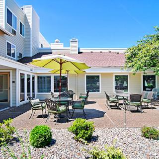 A sunny outdoor patio area with tables, chairs, and an umbrella, surrounded by landscaped greenery.