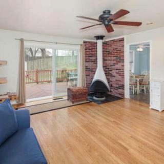 A cozy living room with a central fireplace set against a brick wall, featuring wooden floors and a ceiling fan. Sliding glass doors open up to a patio area.