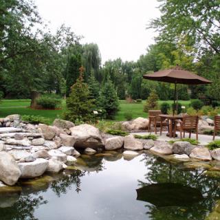 A serene garden scene featuring a tranquil pond surrounded by natural stones and lush greenery, complete with a cozy seating area under an umbrella.