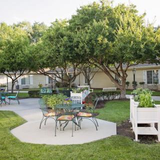 A serene garden with outdoor seating, featuring tables and chairs surrounded by lush greenery and trees.