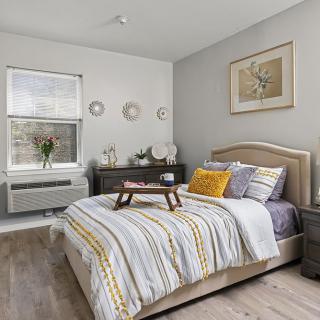A cozy bedroom with a neatly made bed featuring striped bedding, a decorative throw pillow, and a small tray. A dresser is placed against the wall under a window, complemented by framed artwork and decorative plates.
