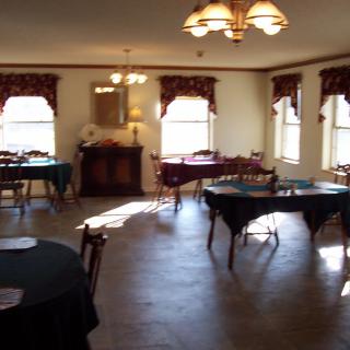 A traditionally decorated dining room with multiple tables and chairs, featuring large windows with curtains and a ceiling chandelier.