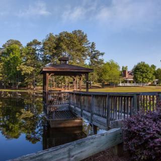 A tranquil scene featuring a gazebo by a pond with a wooden dock extending over the water, surrounded by lush trees.
