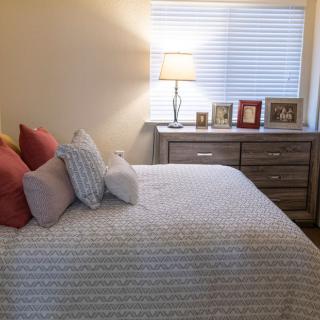 A cozy bedroom with a neatly arranged bed, pillows, and a dresser adorned with photo frames under soft lamp lighting.