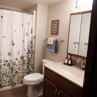 A modern bathroom with a floral shower curtain, white toilet, wooden vanity with a mirror, and neatly arranged towels and toiletries.