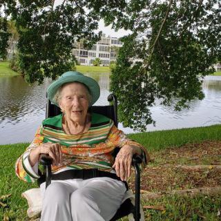 An elderly woman in a wheelchair enjoys a day outdoors by the river, smiling under the shade of trees.