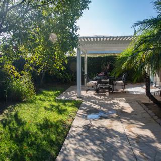 A sunny backyard with a pergola-covered patio and outdoor seating amidst lush greenery.