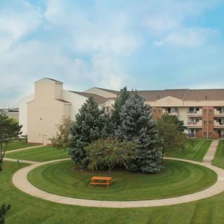 A serene park area with neatly trimmed grass and a circular pathway, surrounded by trees, next to a building under a partly cloudy sky.