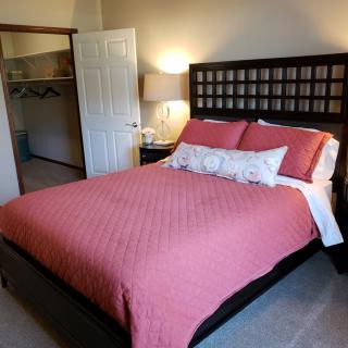 A cozy bedroom with a bed featuring a pink cover and decorative pillows, accompanied by a nightstand with a lamp and an open closet door.
