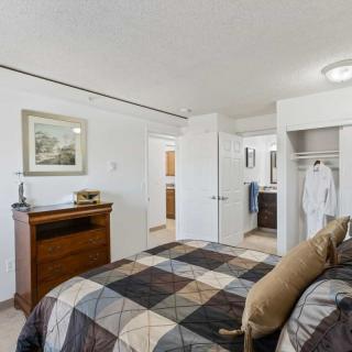 A cozy bedroom featuring a bed with a patterned quilt, a dresser, and an open closet revealing hanging clothes. A bathroom is visible through the open door.
