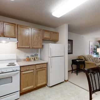 An open-layout kitchen in an apartment, featuring wooden cabinets and essential appliances including a refrigerator and stove.