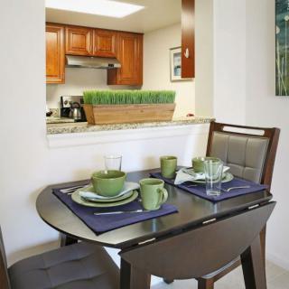 A modern dining area with a wooden table set for two, adjacent to a kitchen with wooden cabinets.
