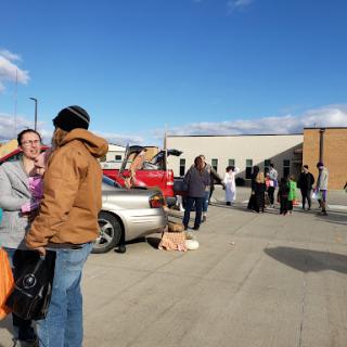 A vibrant community gathering with people and cars in a parking lot on a sunny day.