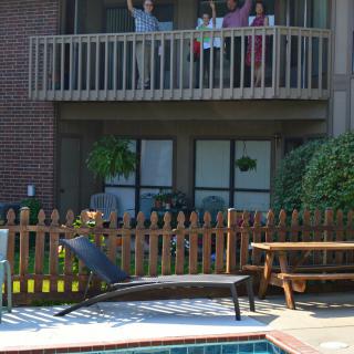 People waving from a balcony overlooking a pool and wooden fence.