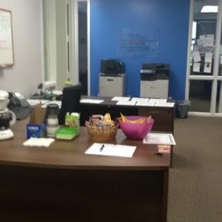 A view of a modern office workspace featuring a desk with various stationery items and printers in the background.