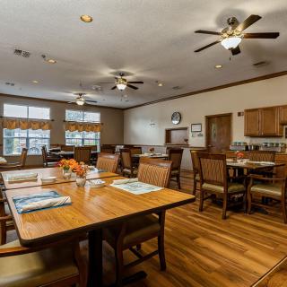 A spacious dining area with wooden flooring, ceiling fans, and a kitchen in the background.