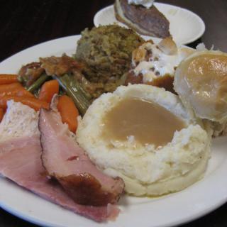 A hearty plate featuring ham, mashed potatoes with gravy, assorted vegetables, stuffing, and a bread roll.