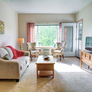 A cozy living room with a sofa, two armchairs, and a coffee table facing a TV, with sunlight streaming through a large window.