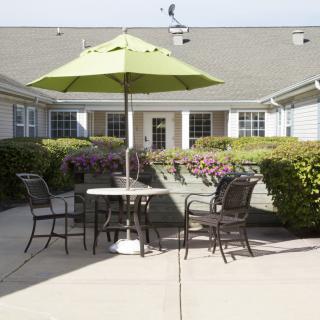 An outdoor patio area with a table, chairs, and a green umbrella surrounded by gardens.